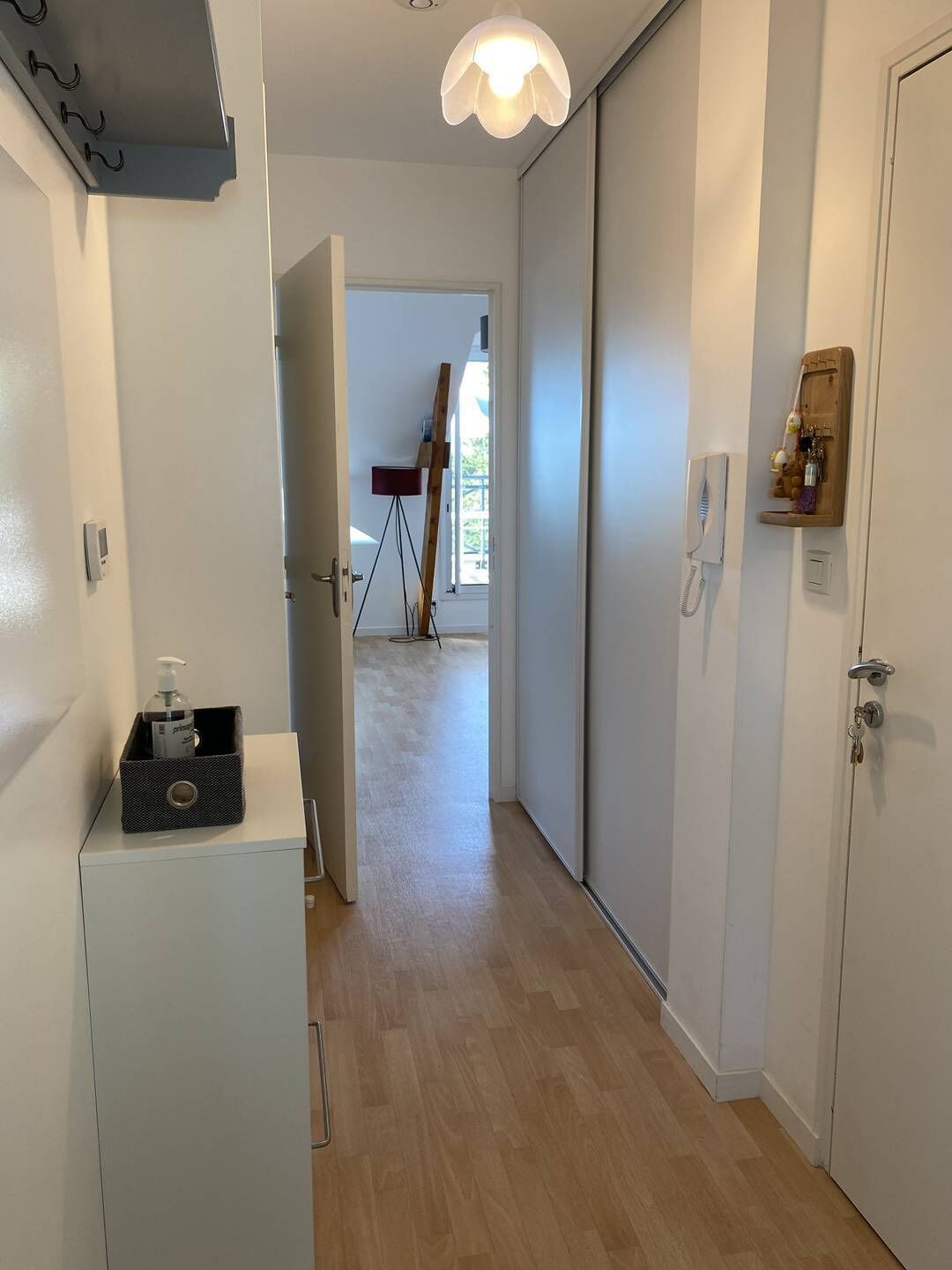 Un couloir avec une armoire blanche, une petite table sur laquelle se trouve une bouteille, et une échelle en bois appuyée contre le mur.