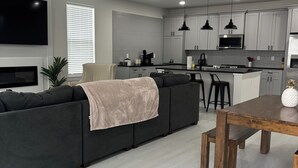 Una sala de estar moderna con un sofá seccional gris, una mesa de comedor de madera y una zona de cocina con armarios grises y un microondas negro.