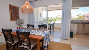 Un coin repas avec une table en bois, une nappe à motifs bleus et blancs, des chaises noires, un ventilateur et une vue sur le balcon et les bâtiments environnants.