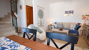 Un salon comprenant un canapé, une table basse et une table à manger avec des chaises. On y trouve également un escalier avec une rampe et une porte.