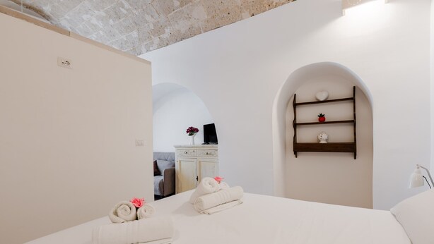 Habitación de hotel con una cama, dos toallas enrolladas, un pequeño estante con adornos y un acceso a otra habitación.
