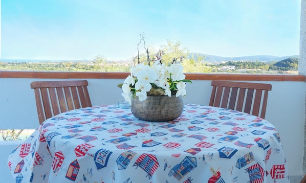 Une table ronde recouverte d’une nappe à motifs, deux chaises en bois et une orchidée blanche en pot sur le balcon.