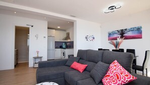 A modern living room with a grey sectional sofa, a small round table, and a dining area with chairs. There is a kitchen in the background with white cabinets and a microwave. - Ref: 262 Beach front apartment with amazing terrace and sea views (Fuengirola)