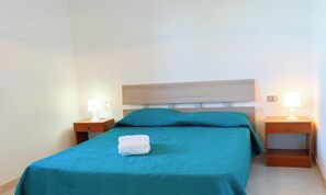 A single bed with a teal bedspread, two bedside tables with lamps, and a white towel on the bed.