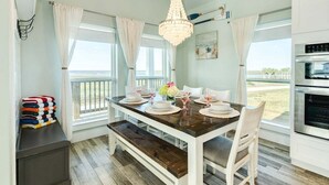 A dining area with a wooden table set for four, white chairs, a chandelier, and a view of the outdoors. - Sunchaser (Galveston)
