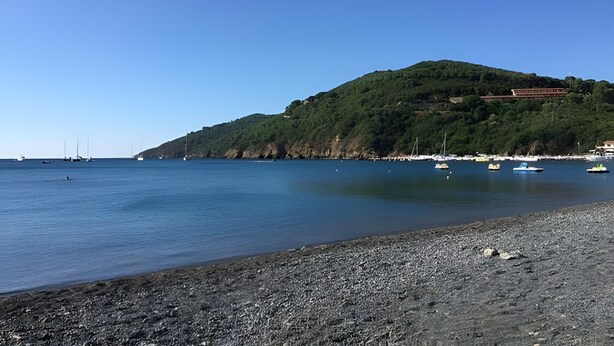 Una playa con barcos, costa rocosa y una colina con edificios.
