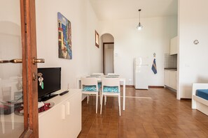 Uma sala de jantar com TV, cadeiras brancas, uma mesa e uma cozinha ao fundo.