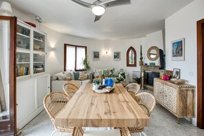 Un coin repas comprenant une table en bois, des chaises en osier, un buffet avec des portes en verre, un canapé et une cheminée.
