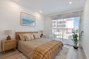 A bedroom with a bed, two bedside tables with lamps, a window with sheer curtains, and a potted plant.