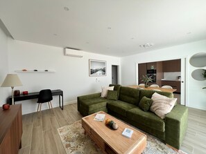 A modern living room with a green sofa, a wooden coffee table, a desk with a lamp, and a kitchen in the background.