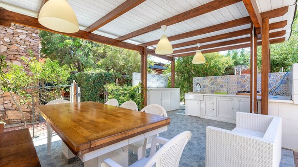 A covered outdoor dining area with a wooden table and white wicker chairs, stone walls, and a tiled backsplash in the background. - Apartment 'Casa 20' with Private Terrace, Wi-Fi and Air Conditioning (Stintino)
