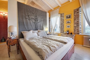 Un dormitorio con una cama grande, una mesita de noche, un tapiz en la pared, una ventana con cortinas y una pared de piedra.