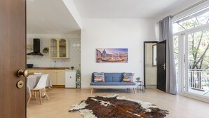 Een moderne woonkamer met een kussen van runderleer, een bank met kleurrijke kussens, een eethoek met een tafel en stoelen, een keuken met kastjes en een fornuis, en een groot raam met uitzicht op gebouwen.
