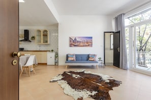 Uma sala de estar moderna com tapete de couro, sofá com almofadas coloridas, área de jantar com mesa e cadeiras, cozinha com armários e fogão, e uma grande janela com vista para edifícios.