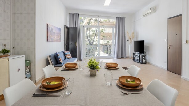 Een eethoek met een tafel voor vier personen, een bank, een televisie en uitzicht op een balkon.