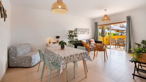 A dining area with a table, chairs, and a sofa. There are plants on the table and a painting on the wall. - Apartment 'Royal Cabanas Golf' with Shared Pool, Wi-Fi and Air Conditioning (Conceição de Tavira)