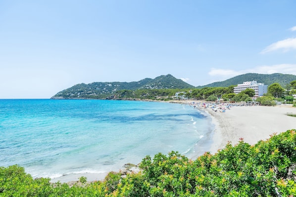 Una playa costera con agua turquesa cristalina, una orilla arenosa y colinas verdes al fondo.