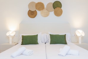 Cama doble con ropa de cama blanca, dos almohadas verdes y toallas blancas enrolladas. Hay dos mesitas de noche con lámparas.