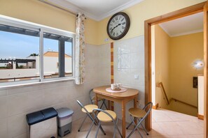 A small room with a window, a clock, a table with chairs, a trash bin, and a door leading to another room.