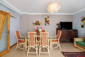 A dining area with a glass table, wicker chairs, a chandelier, artwork on the wall, a television, and a sofa.