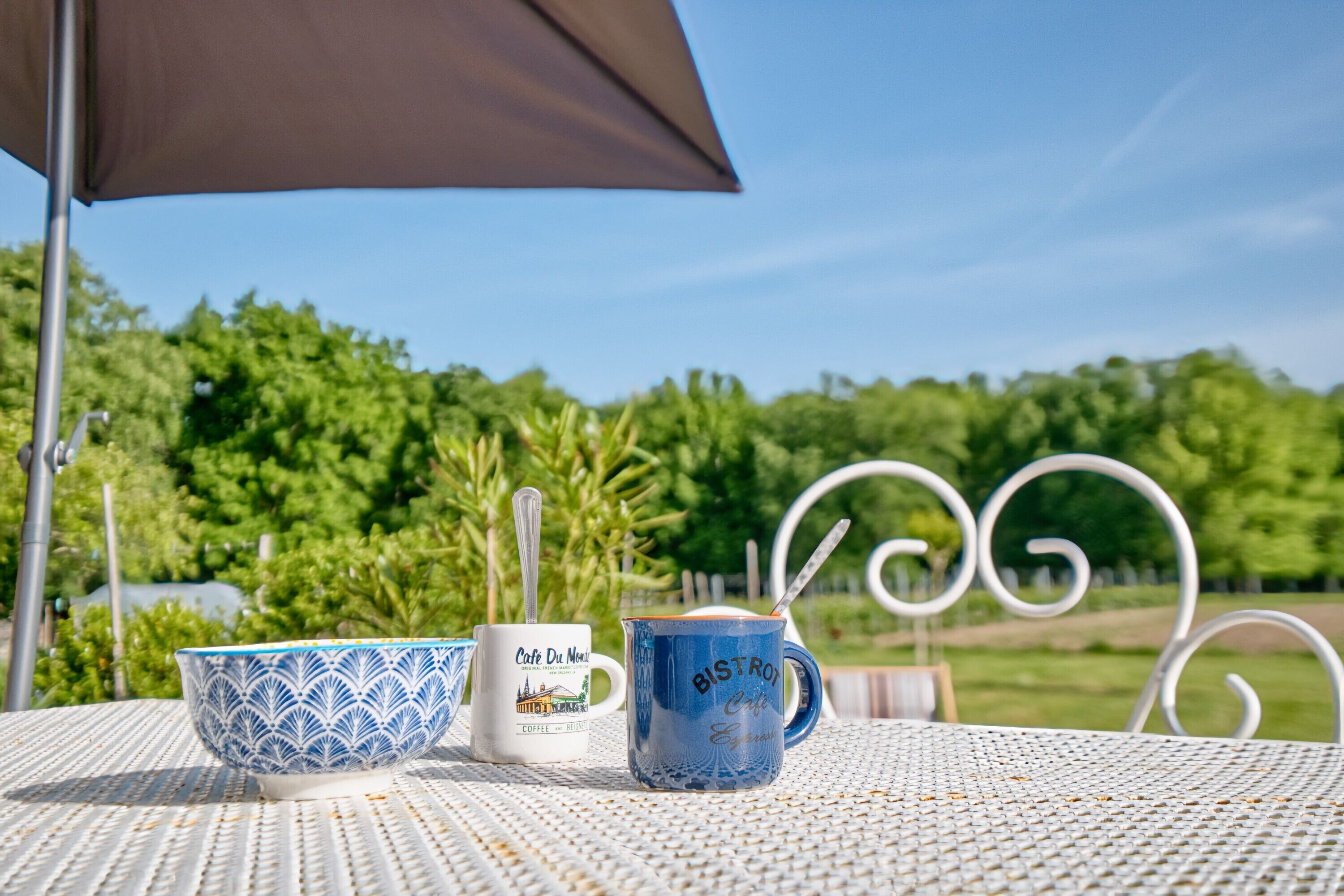 A table with two cups and a spoon, a brown umbrella, and a metal chair with a decorative backrest.