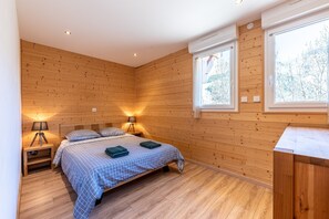 Un dormitorio con piso de madera, una cama con cubrecama cuadriculado, dos lámparas de noche y una ventana con vista a árboles.