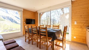 A dining area with a table set for four, wooden chairs, a TV mounted on the wall, and large windows offering a mountain view.