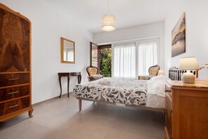 Une chambre à coucher comprenant un lit, une commode, une chaise, une table, un luminaire suspendu et une fenêtre avec des rideaux.