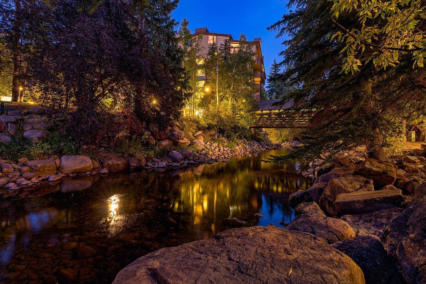 Une vue nocturne d’une rivière bordée de rochers et d’arbres, d’un pont en bois et d’un bâtiment illuminé.