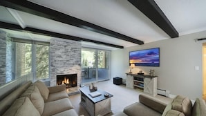 A modern living room with a fireplace, large windows, and a flat-screen TV mounted on the wall.