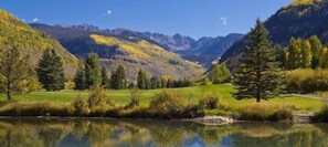 Un paysage paisible avec un lac dans lequel se reflètent les arbres et les montagnes, une zone herbeuse et un ciel dégagé.