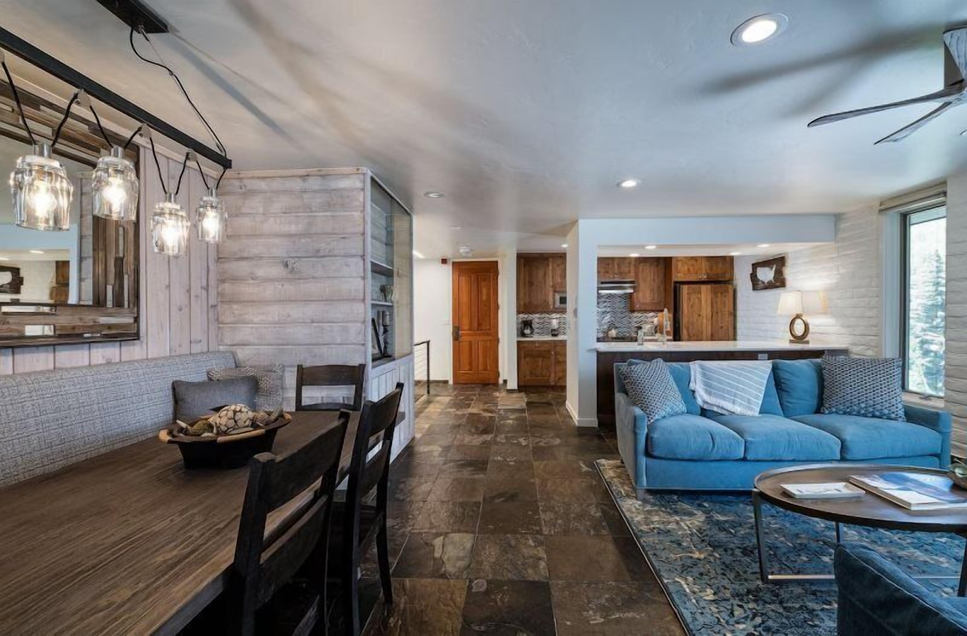 Une cuisine moderne avec des meubles en bois et un îlot central, un coin repas avec une table et des chaises en bois, et un salon avec un canapé bleu et une table basse.