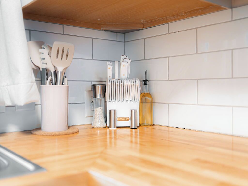 Una cocina con azulejos blancos, una encimera de madera y varios utensilios, incluyendo espátulas y cuchillos.