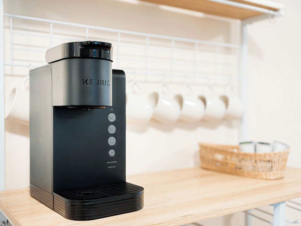 Eine schwarze Keurig-Kaffeemaschine auf einem Holztisch, im Hintergrund eine Korb mit Kaffeekapseln.