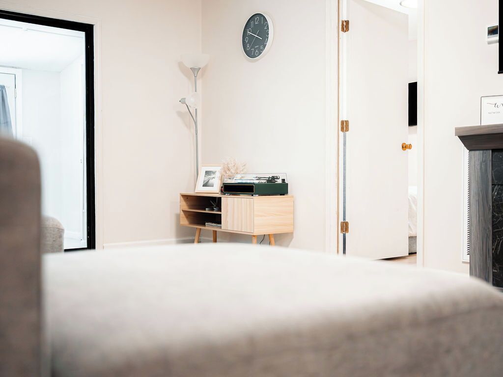 Una habitación minimalista con una cama, una pequeña mesita de madera con un reloj, y una puerta que da a otra habitación.