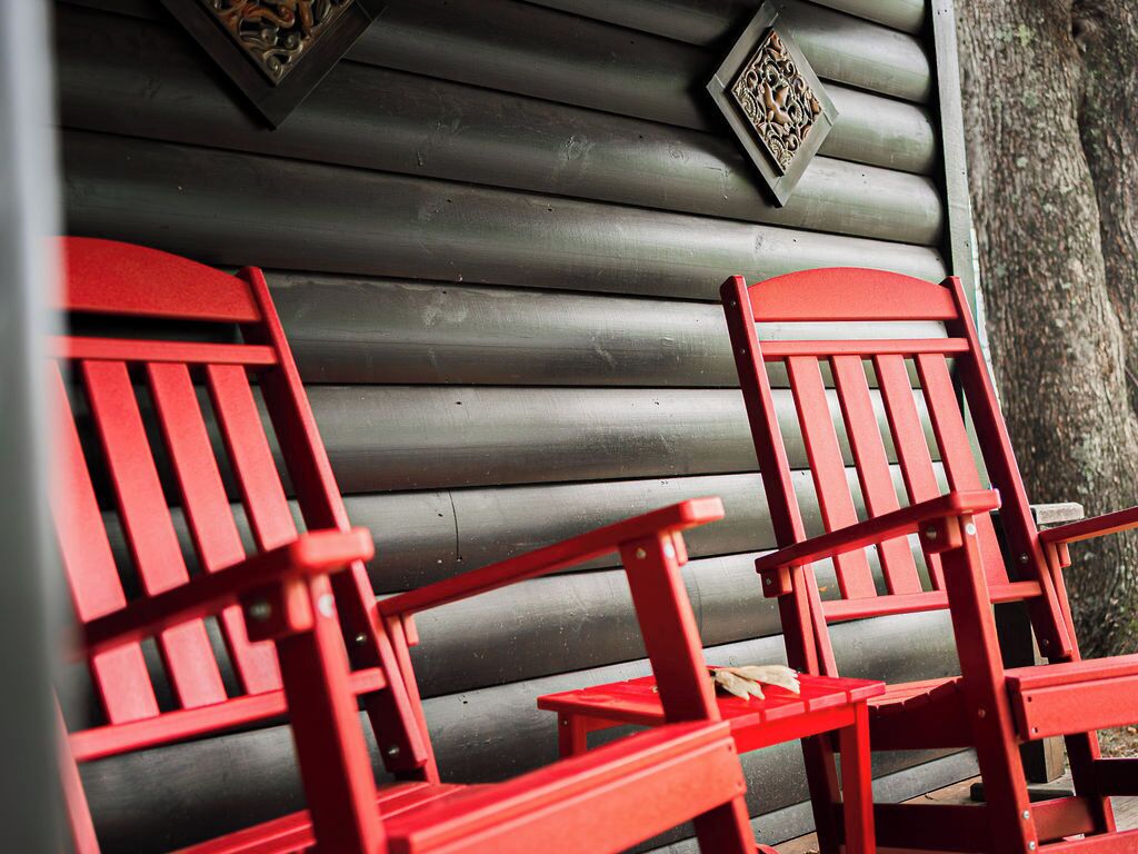 Two red Adirondack chairs on a porch with a wooden wall and decorative metal elements.