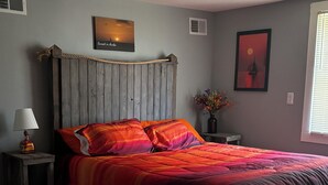 Une chambre à coucher avec une tête de lit en bois, un lit recouvert d’une courtepointe colorée, une lampe de chevet et deux œuvres d’art accrochées au mur.