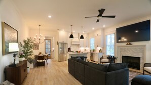 A modern kitchen with white cabinets, a dining area with a table and chairs, a living room with a sofa and ottoman, and a fireplace.