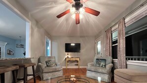 Una sala de estar con una mesa de centro de vidrio, dos sillones, un televisor y un ventilador de techo.