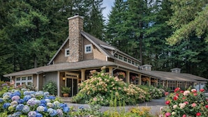 A large, two-story house with a stone chimney, surrounded by lush greenery and vibrant flowers.