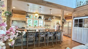 A kitchen with a central island, wooden bar stools, hanging plants, and a stainless steel refrigerator.