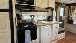 A compact RV kitchen with a stove, microwave, sink, and white cabinetry. - Montana Moon on the Lake "Outback" RV #7 (Babb)