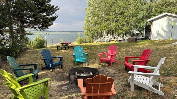 A grassy outdoor area with several colorful Adirondack chairs arranged around a fire pit. A small shed and a table with chairs are visible in the background. - Montana Moon on the Lake "Outback" RV #7 (Babb)