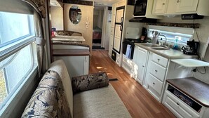 A compact RV interior with a kitchenette, a seating area with a patterned cushion, and a small bathroom with a mirror. - Montana Moon on the Lake "Outback" RV #7 (Babb)