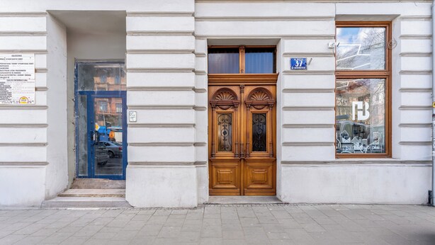 Fachada de edificio con un gran puerta doble de madera, una ventana pequeña con un letrero, y una puerta azul con un letrero.