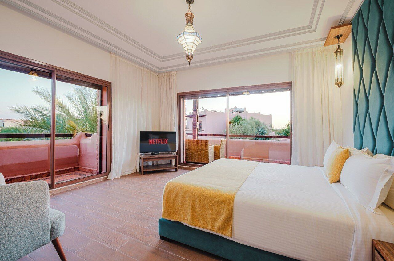 A hotel room with a large bed, a TV on a stand, a balcony with a sofa, and a view of the outside.
