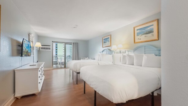 Ein Hotelzimmer mit zwei Betten, einem Schreibtisch, einem Stuhl, einem Fernseher und einem Balkon mit Blick auf Gebäude.