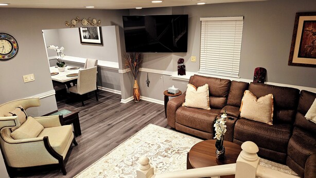 A modern living room with a brown sofa, beige armchairs, a dining area with a white table, and a TV mounted on the wall.