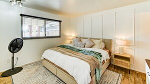 Une chambre à coucher comprenant un lit, des lampes de chevet, un ventilateur sur pied, une fenêtre avec des stores et un luminaire au plafond.