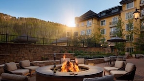 A circular outdoor seating area with a fire pit, surrounded by wicker chairs and tables, set against a backdrop of a hotel building and a forested hillside. - The Ritz-carlton Club, Vail 4 Bedroom Luxury Residence (Vail)